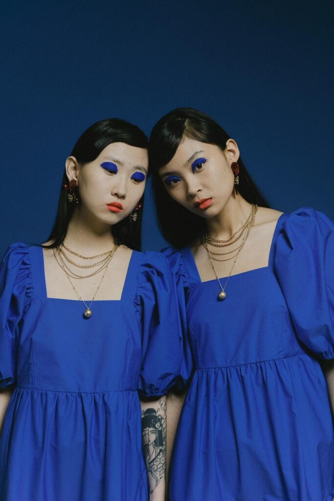 Two models in matching blue dresses with striking blue eyeshadow, posing against a blue backdrop.